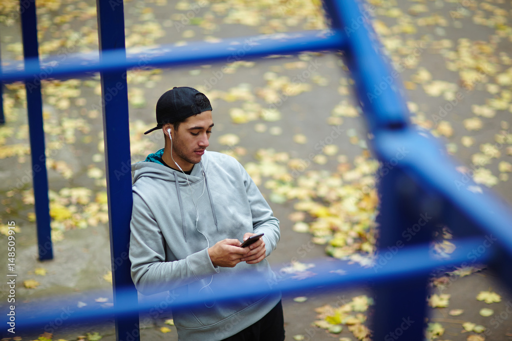 High angle view of handsome bearded man in sportswear standing at horizontal bar while listening to music on smartphone, portrait shot