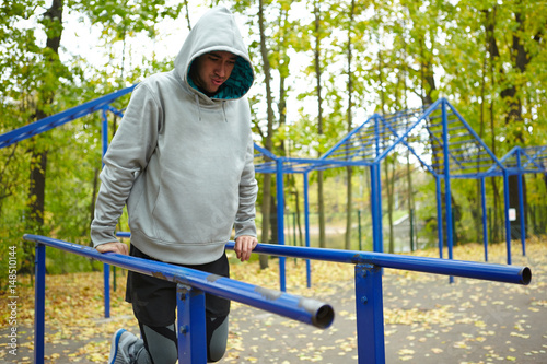 Young sportsman in hoodie doing street workout exercise, legs bent at knee, full-length portrait