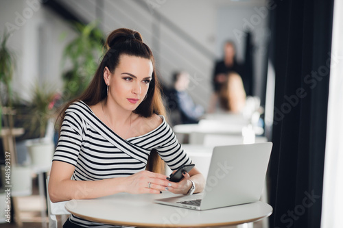 Texting woman sitting at the table and holding the mobile phone, thinking. Adult female sitting with a laptop and looking away.