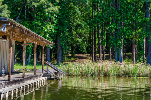 Lake boat house resting on reflective lake shoreline water.  Relaxing tranquil outdoor scene of woods, grass and water with boat house.  