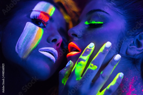 Sexy lesbian fashion models kissing in uv neon light with fluorescent glowing Body Art make-up . Low key dark image. Soft focus image.