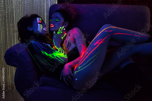 Sexy lesbian fashion models in uv neon light with fluorescent glowing Body Art make-up lying and kissing. Low key dark image. Soft focus image. photo