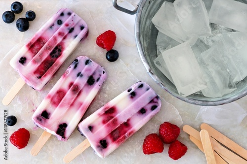 Group of mixed berry yogurt popsicles with fruit and ice pail on a white marble background photo