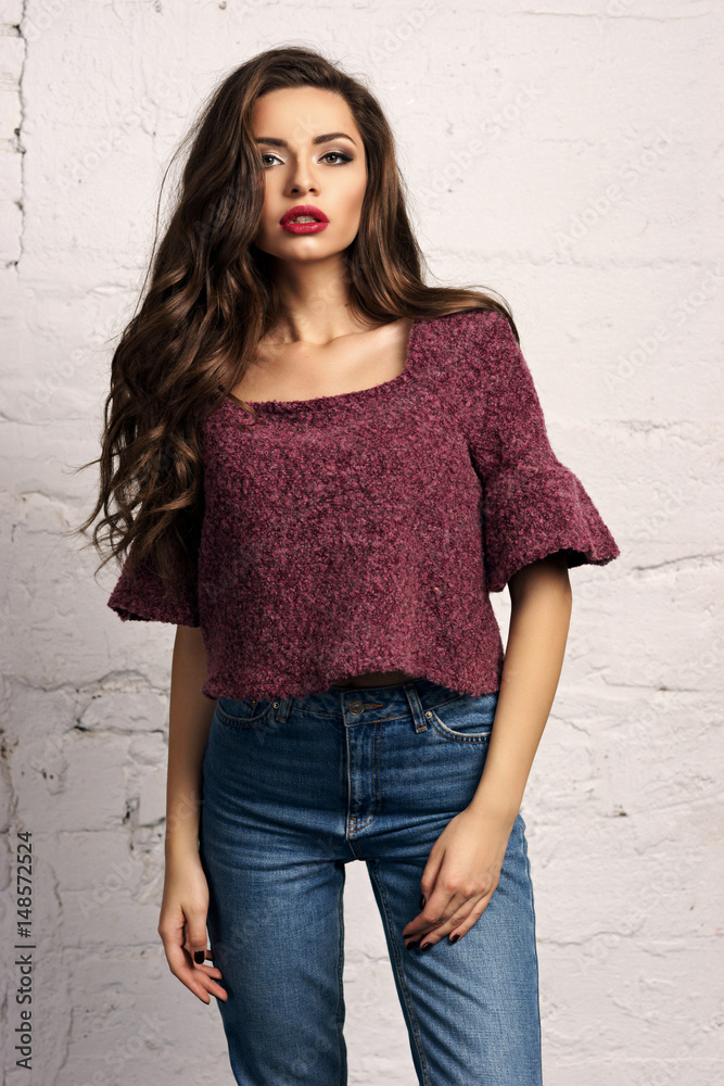 Young beautiful gorgeous female model in blue jeans and purple pullover posing against white brick wall. Stunning glamorous girl with long curly hair