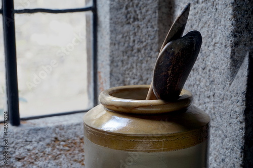 Old wooden cooking spoons in a large ceramic jug or pot, with stone walls and window in background Organic