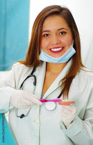 Beautiful woman doctor posing for the camera using a surgery mask in her chin while she is brushing a fake dental plaque photo