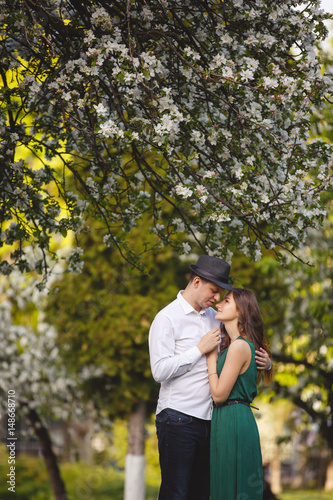 Young man and woman couple in a blooming apple garden. Tender holding each other. Spring lovestory. Brown-haired girl with long hairs. Young family