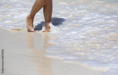 Vacation concept image. A caucasian woman is walking barefoot in the water line. Foam on white sand.