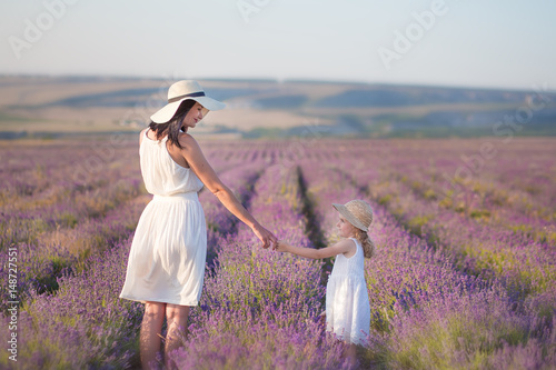 Young beautiful lady mother with lovely daughter walking on the lavender field on a weekend day in wonderful dresses and hats.