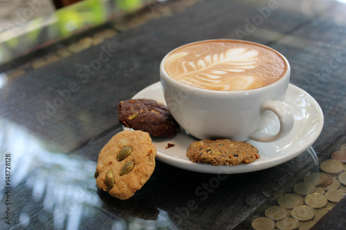 Latte coffee with froth flower art and biscuits on the saucer served outdoors in sunlight on a glass table with inlaid coins