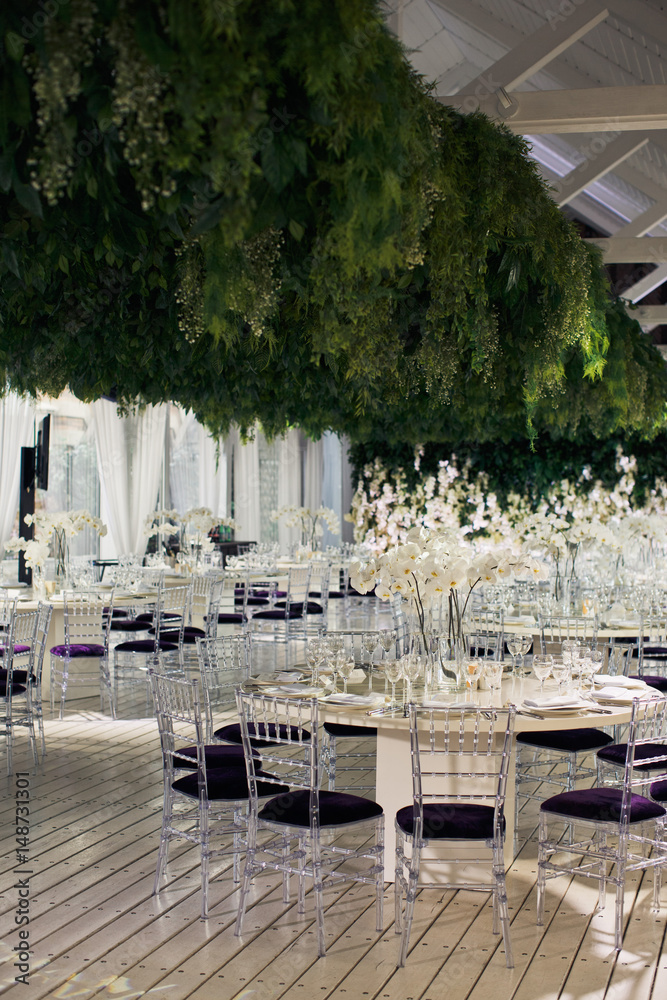 Violet chairs stand at white dinner tables under large festoon of greenery