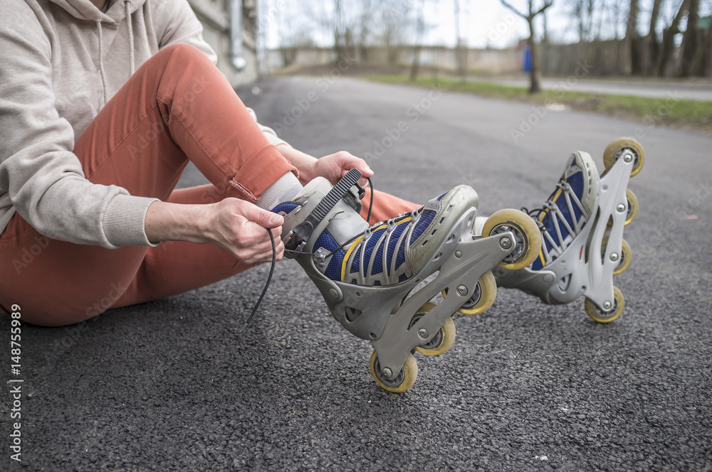 Obraz premium Girl sitting on the road and tying the laces on roller skates, close-up. Concept: sports, healthy lifestyle.