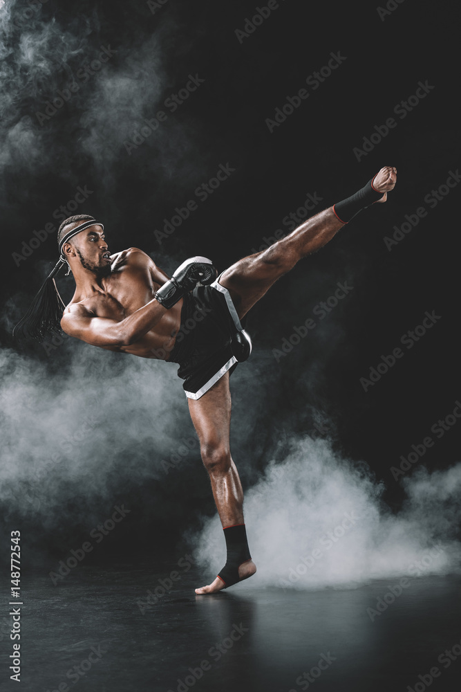 Side view of concentrated Muay Thai fighter practicing kick isolated on black, fight club concept