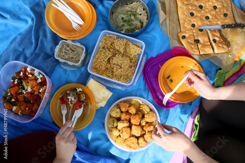 Unrecognizable people eating various picnic food: roasted vegetables salad, baba ghanoush, gluten-free crackers, foccacia bread, gluten-free and sugarfree dates cake. Top view. 