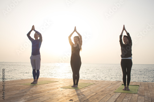 group of people in yoga poses stand on the wooden pier, sunrise in the sea background 