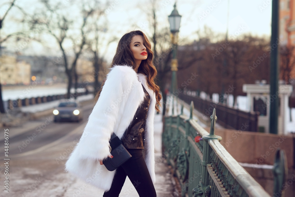Young beautiful stylish woman walking down the street on a cold winter snowy day. Fashionable girl wearing black blouse and trousers and white fur coat. Female with long curly hair. - obrazy, fototapety, plakaty 