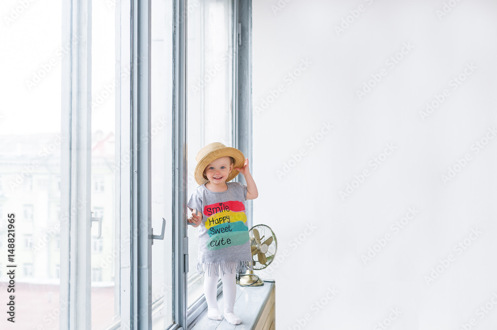 Baby girl in a straw hat and a beautiful children's shirt costs on a window sill.