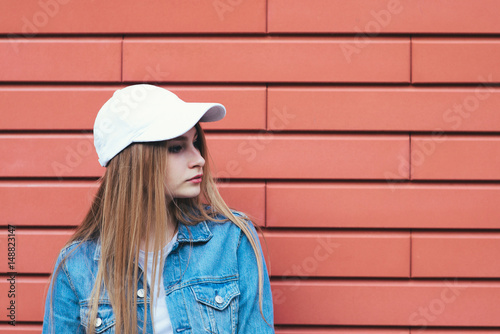 Portrait of stylish hipster girl in a cap and denim jacket matched looks toward the street on a bright background. Girl on a background of the red wall © bodnarphoto