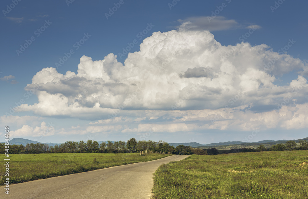 Summer countryside landscape with stormy cumulonimbus clouds in the sky and empty road crossing the grasslands of the Transylvania region, Romania.