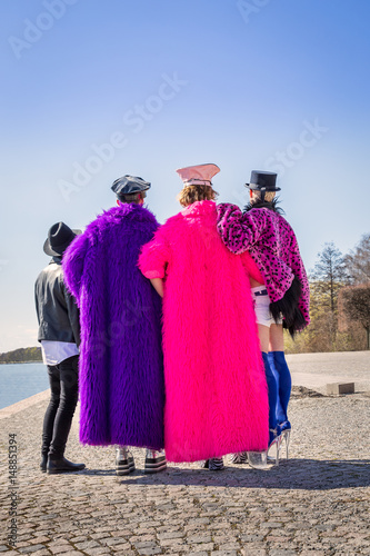Caucasian men in colorful fake furs and hats standing on a city quay together and holding each other, seen from behind.   © Pebo
