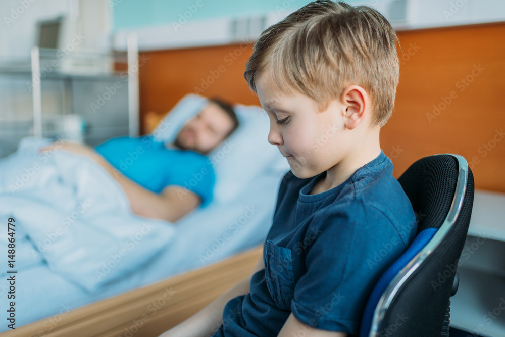 little son sitting on chair near his sick father sleeping on hospital bed at ward, dad and son little son sitting on chair near his sick father sleeping on hospital bed at ward, dad and son