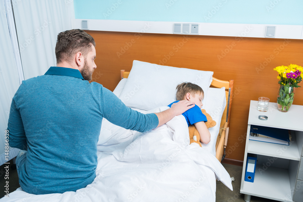 High angle view of father touching sick little son lying in hospital bed with teddy bear, dad and son in hospital High angle view of father touching sick little son lying in hospital bed with teddy bear, dad and son in hospital