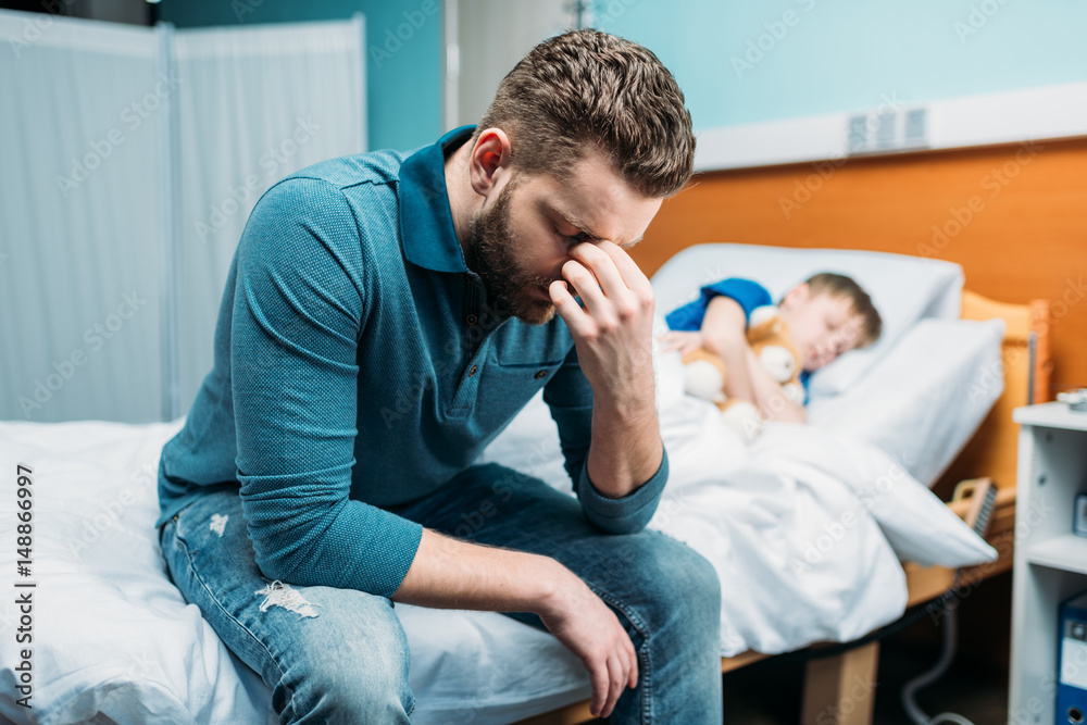 side view of tired dad sitting near sick son in hospital bed side view of tired dad sitting near sick son in hospital bed