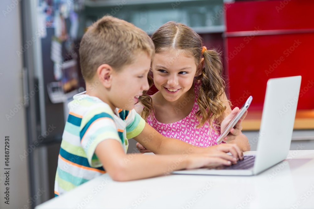 Siblings using laptop and mobile phone in kitchen Siblings using laptop and mobile phone in kitchen