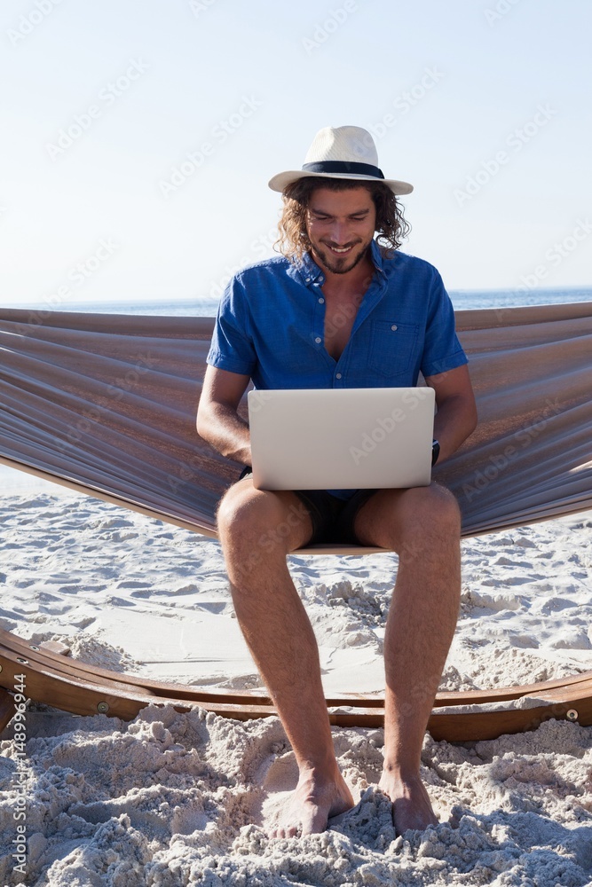 Man using laptop while sitting on hammock at beach