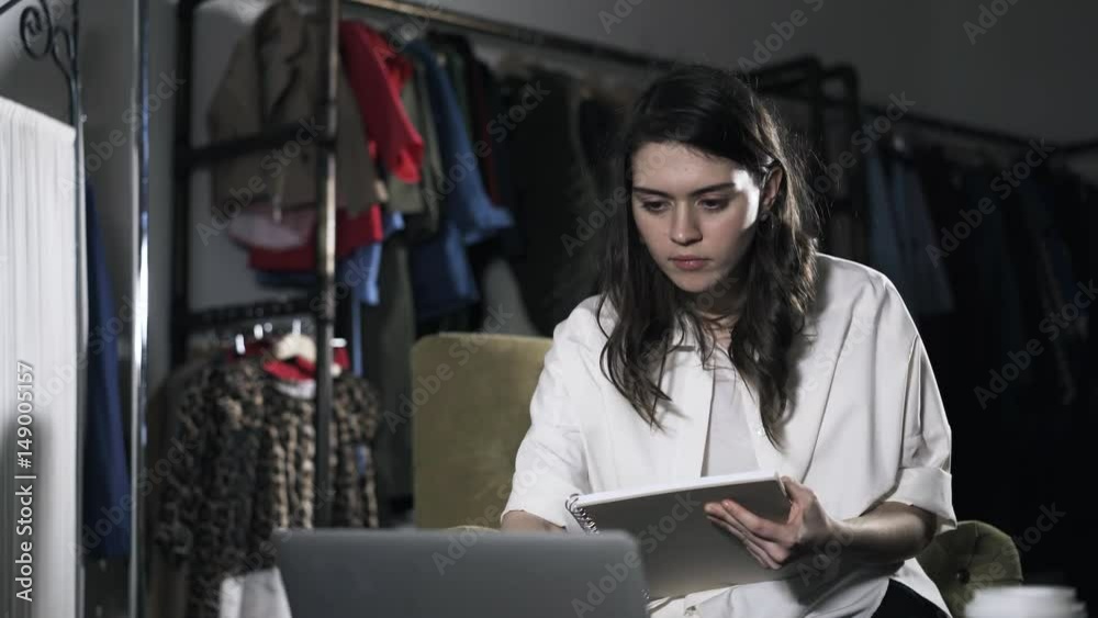 Young woman is looking at her laptop screen and writing in a notebook sitting in an armchair at home. Locked down real time medium shot Young woman is looking at her laptop screen and writing in a notebook sitting in an armchair at home. Locked down real time medium shot