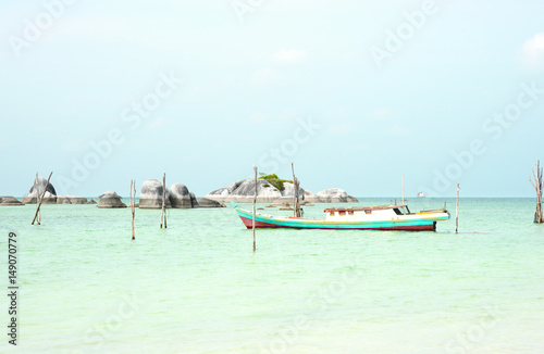 Empty fishing boat docked at the turquoise colored ocean coast near white sand beach in the afternoon with rocks on the ocean horizon in Belitung, Indonesia