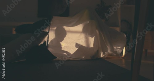 Silhouette of cute girl reading a book to his little brother inside a blanket fort in the evening, lit by a lamp from inside. 4K UHD RAW edited footage