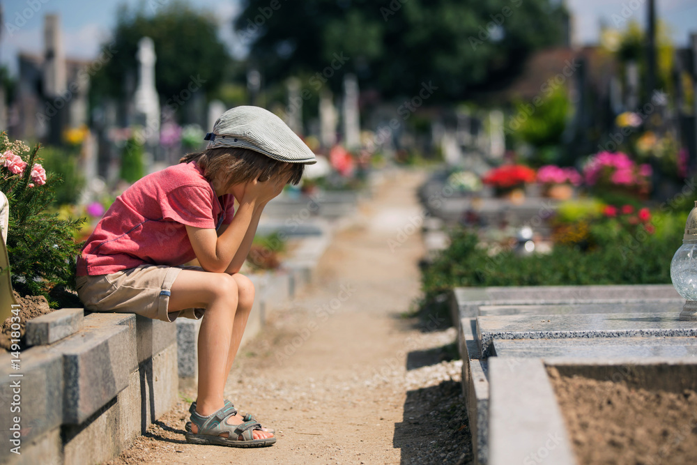 Sad little boy, sitting on a grave in a cemetery, feeling sad