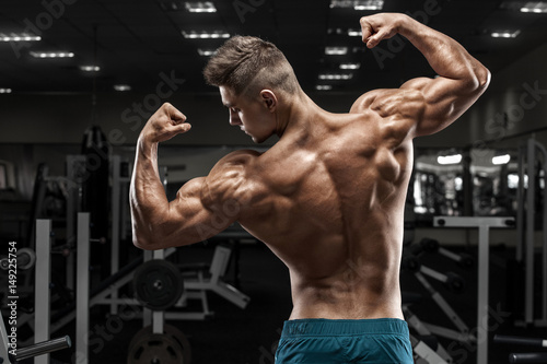 Rear view muscular man posing in gym, showing back and biceps. Strong male naked torso, working out