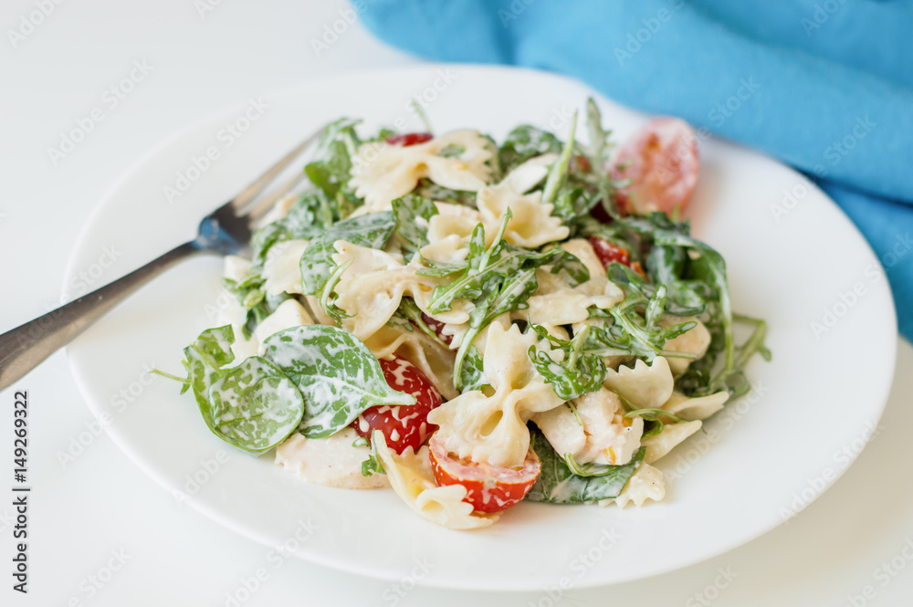 Portion of pasta salad with rucola leave, baby spinach, farfalle, sliced cherry tomatoes and chicken meat dressed with white sauce and served on white plate with fork and blue napkin Portion of pasta salad with rucola leave, baby spinach, farfalle, sliced cherry tomatoes and chicken meat dressed with white sauce and served on white plate with fork and blue napkin