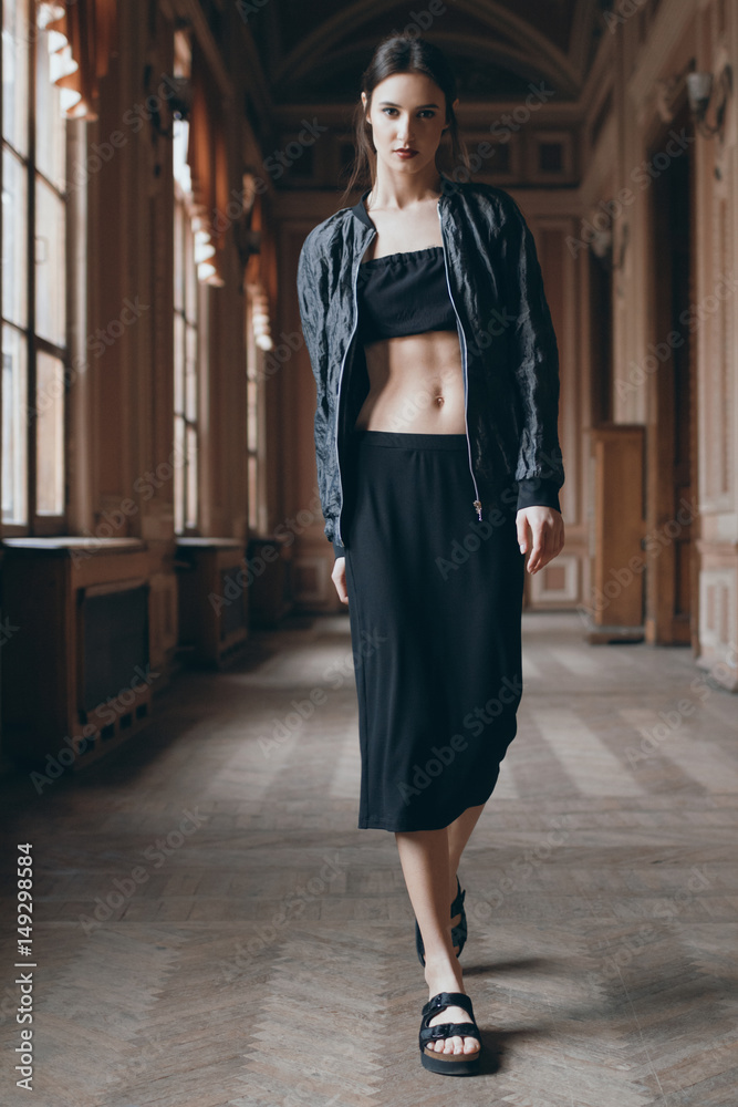 Beautiful model in black top, windbreakert, skirt and slippers looking at  the camera and walking in theatre corridor. Brunette romantic girl with  sexy belly in classic museum interior. Elf hairstyle Stock Photo