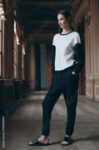 Brunette glad girl dreaming in classic museum interior. Fashion lookbook. Attractive romantic teen model in sweater, trousers and slippers looking out the window with hand in pocket in long corridor.