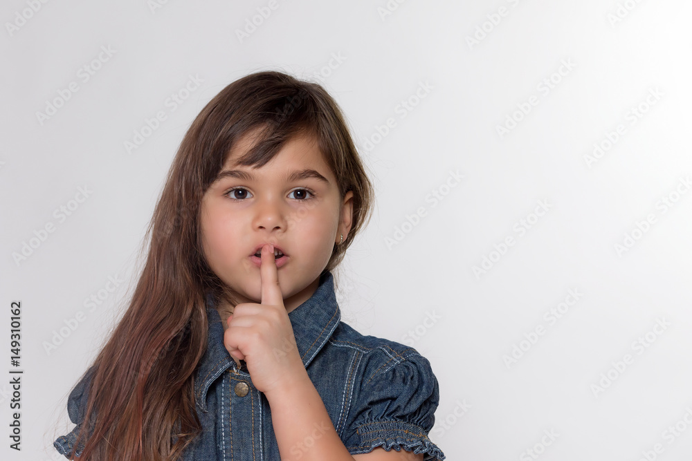 Portrait of brunette long haired little girl gesturing shush. Girl is facing the camera on a gray background. Portrait of brunette long haired little girl gesturing shush. Girl is facing the camera on a gray background.
