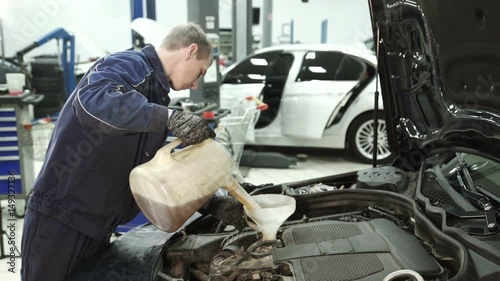 Master Service Center pours oil from a watering can to the motor car. Young mechanic in uniform and gloves through a makeshift funnel replaces technical liquid over the hood. Man of European