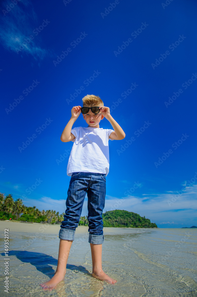 Adorable boy having fun on the tropical beach. White t-shirt, dark trousers and sunglasses. Barefoot on white sand