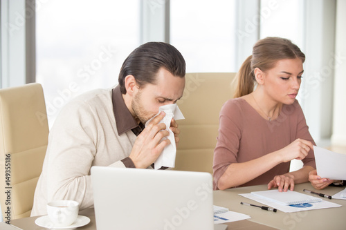 Sick man with handkerchief sneezing blowing nose while working with coworkers, businessman caught cold, seasonal flu. Pandemic influenza, disease prevention, air conditioning in office cause sickness