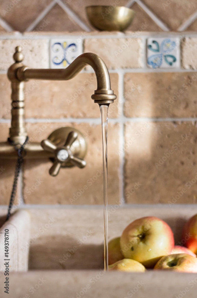 Brass faucet at the vintage granite sink. Running water is washing apples  in the sink. There is also small brass bowl on the top of stone shelf.  Stock Photo | Adobe Stock