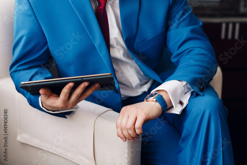 Businessman keep a digital tablet in hand whilst sitting on a sofa in a blue suit. on hand expensive mechanical watch with leather strap. shirt with cufflinks