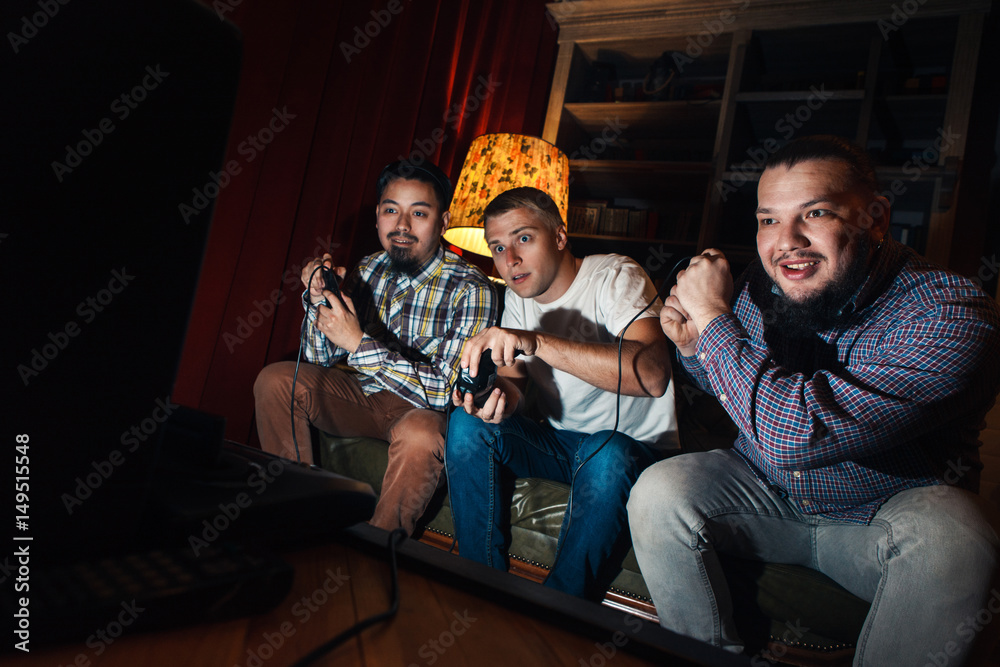 Three excited and concentrated young guys with joysticks play video game. Tensioned youth sit on couch in dark room with gamepad.