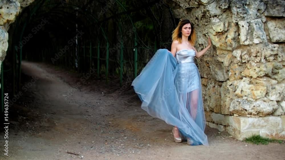 Skinny girl in silver and blue dress in high heeled shoes stands near stony wall of arc and dances as gipsy preparing to pose during photo shoot in antique estate. Skinny girl in silver and blue dress in high heeled shoes stands near stony wall of arc and dances as gipsy preparing to pose during photo shoot in antique estate.