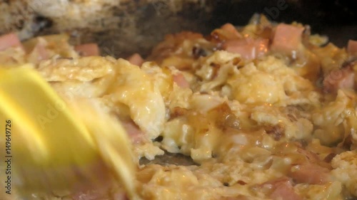 Woman's hand stirs fried sausages, onions and egg mixture in a frying pan with a yellow silicon scapula. Close-up. 50 fps