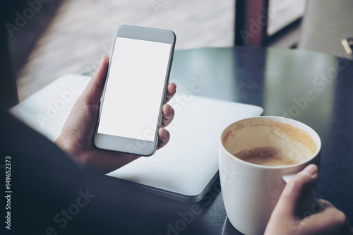 Mockup image of business woman holding mobile phone with blank white screen with latop and coffee cup on wooden table in cafe