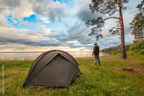 Tourist near camp tent on lake shore, enjoying the view at sunset