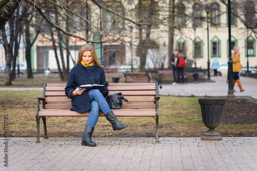 young european woman with blue coat is reading some book while she sitting on bench in park at autumn time