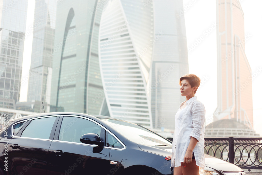 Beautiful young woman standing near black car on skyscrapers background